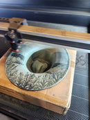 Decorative wooden box with a glass lid featuring engraved patterns, held open by a tool.